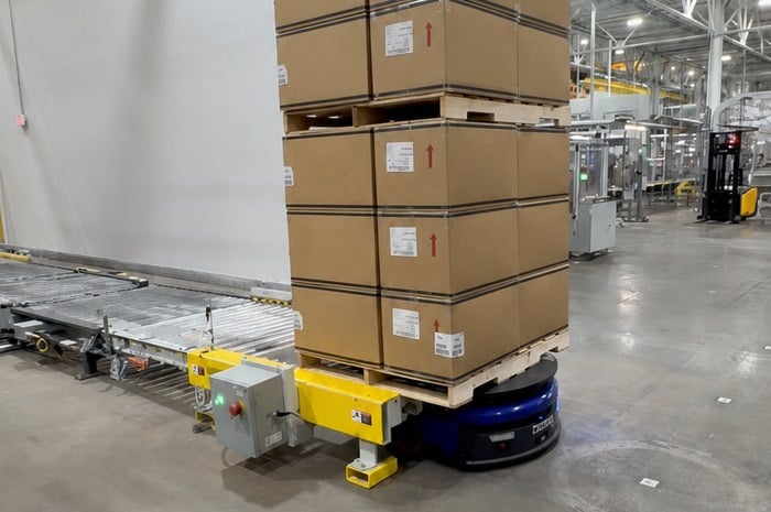 Prime factory AMR robot docked at conveyor transfer station with full pallet — point-to-point drop-off in live manufacturing facility