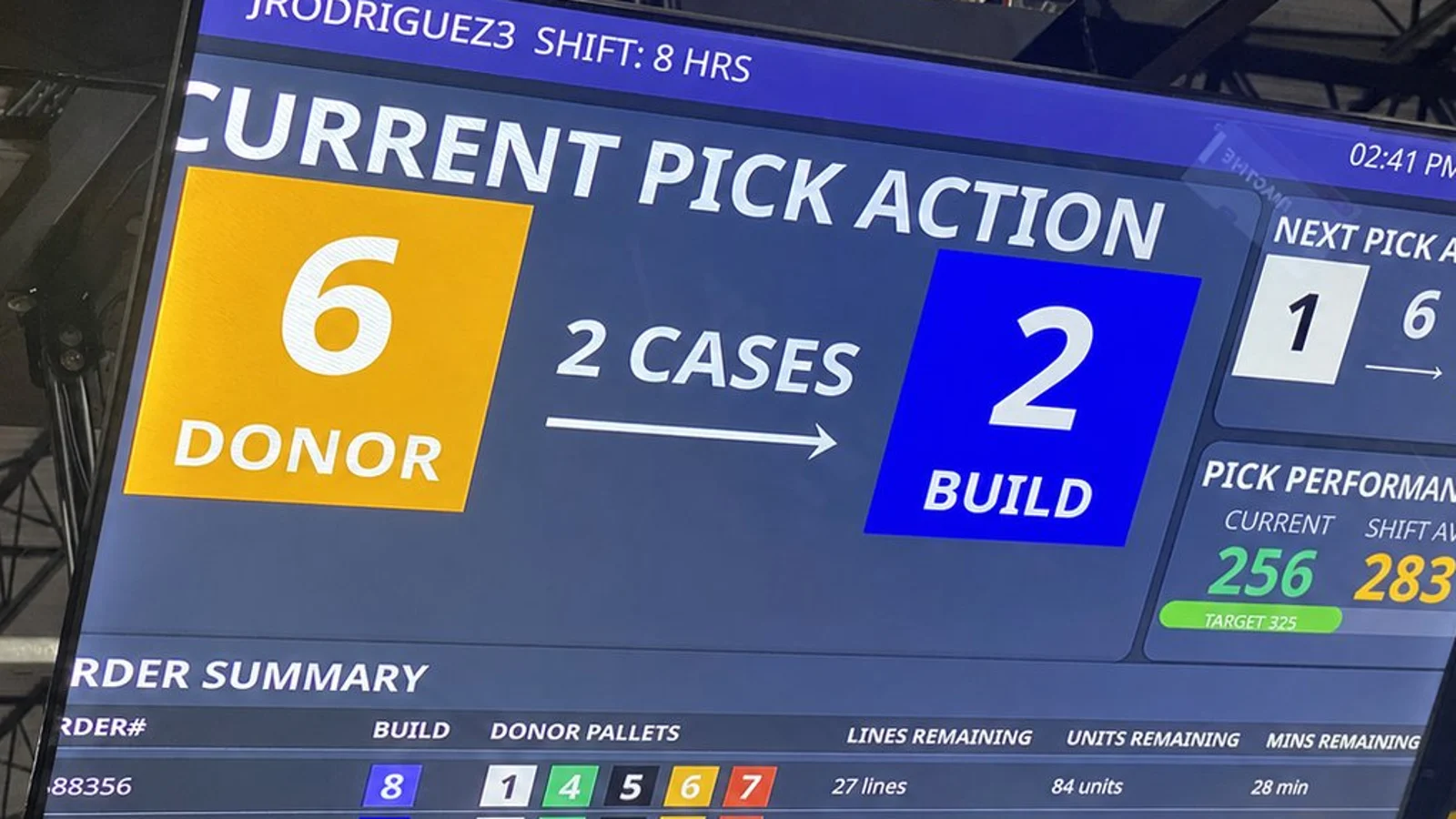 The overhead operator display at a live Tall Pallet Station, showing a single clear instruction: pick 2 cases from donor pallet 6, build to pallet 2, with next pick, pick performance, and order summary alongside